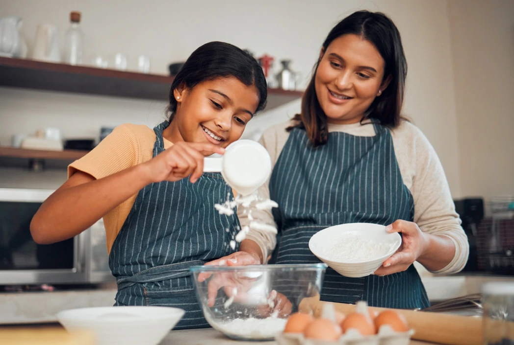 A smiling woman and young girl, both in striped aprons, joyfully baking. The girl pours flour into a mixing bowl on a kitchen counter with eggs.