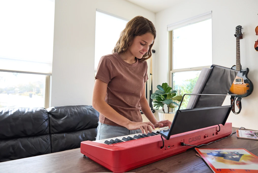 Una mujer joven con una camisa marrón toca un teclado Casio rojo en una habitación luminosa. Las guitarras cuelgan de la pared, y una planta está junto a la ventana, creando un ambiente relajado.