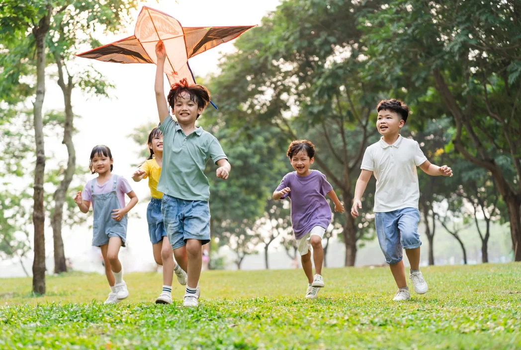 Five joyful children are running through a grassy park, with one boy leading, holding a kite above his head. The scene is lively and carefree.
