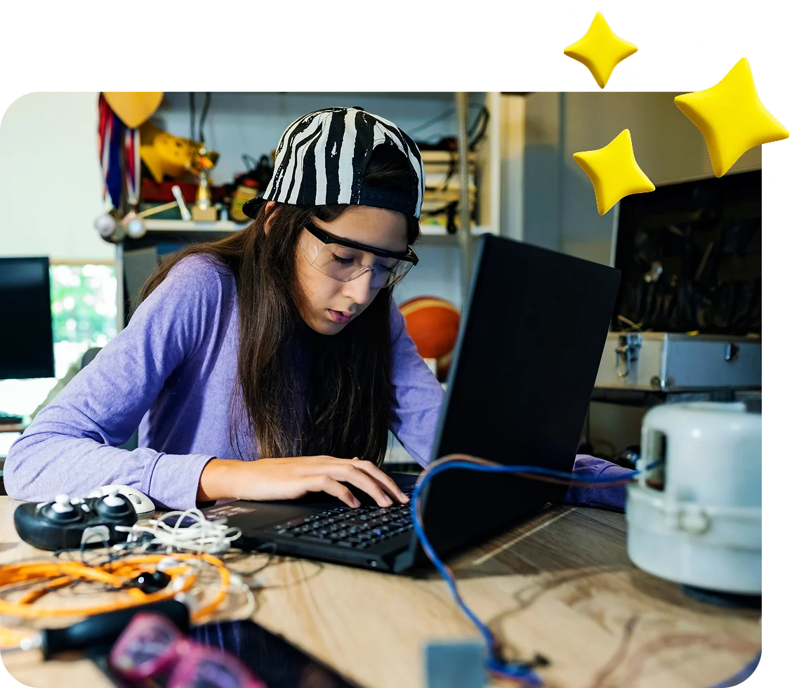 Learner wearing safety glasses works on a laptop at a table with electronic components, wires, and devices. Decorative yellow star graphics appear in the upper corner, highlighting a technology-focused learning environment.