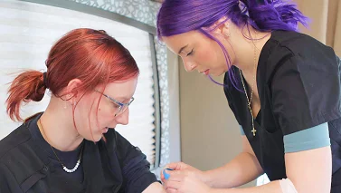 Two healthcare workers in scrubs, one with purple hair, focus on a medical procedure. The scene conveys professionalism and care.