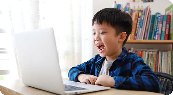 A young boy, smiling widely, sits at a desk using a laptop. He looks engaged and excited. Bookshelves filled with colorful books are in the background.
