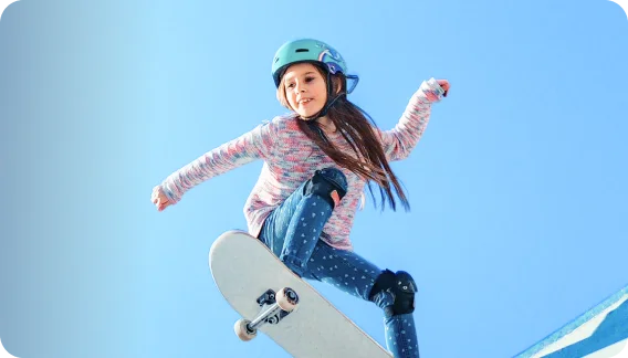 A young girl in a blue helmet performs a skateboard trick against a clear blue sky, wearing a colorful sweater and knee pads.