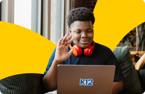 Person seated indoors using a laptop with a K12 logo, wearing red headphones around the neck and raising a hand during an online learning session. Yellow graphic shapes appear in the background.