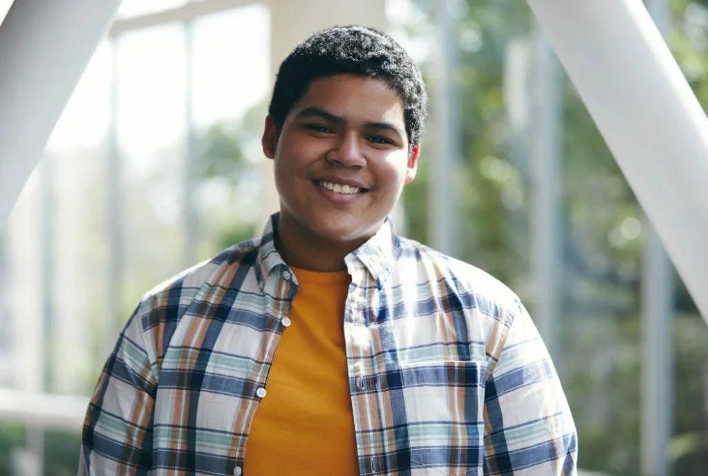 A smiling young man in a yellow shirt and open plaid overshirt stands in a bright, sunlit setting, conveying warmth and confidence.
