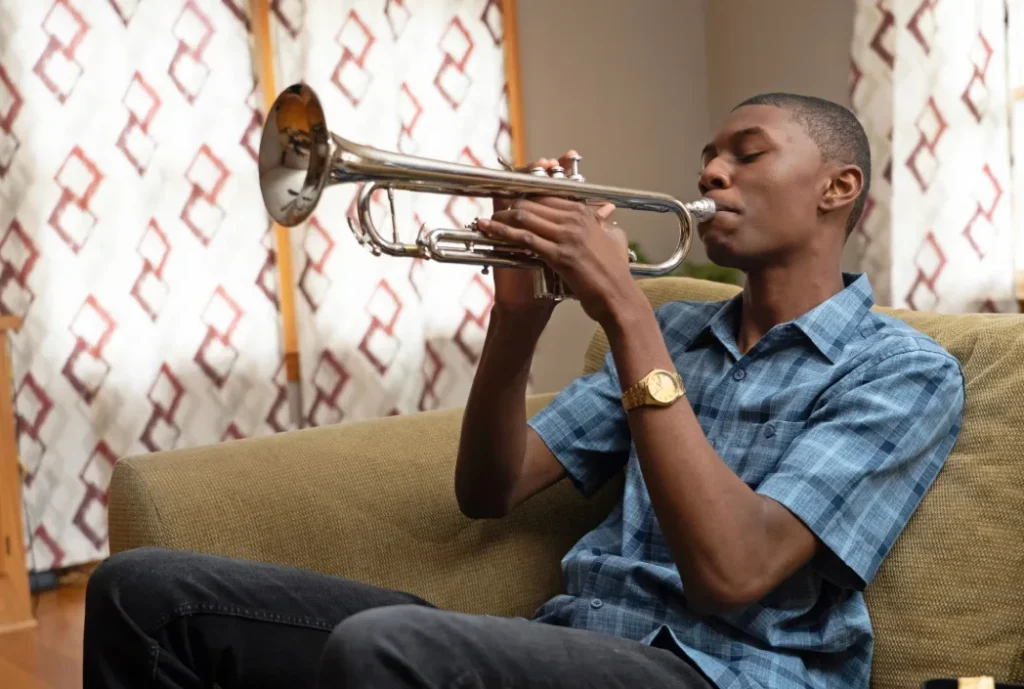 A young man in a blue plaid shirt sits on a couch, eyes closed as he plays a silver trumpet, creating a calm, intimate scene.